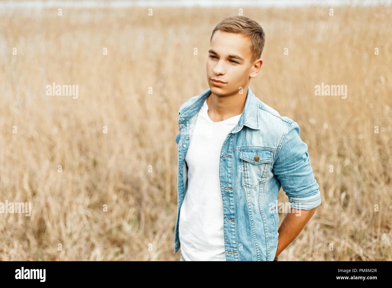 Handsome stylish young man in a jeans jacket and a white shirt in nature in a gray grass Stock Photo