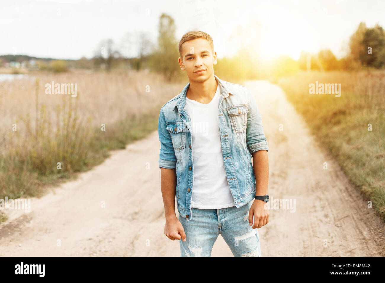 Handsome young man in a jeans jacket and a white T-shirt with torn jeans in the nature at sunset Stock Photo