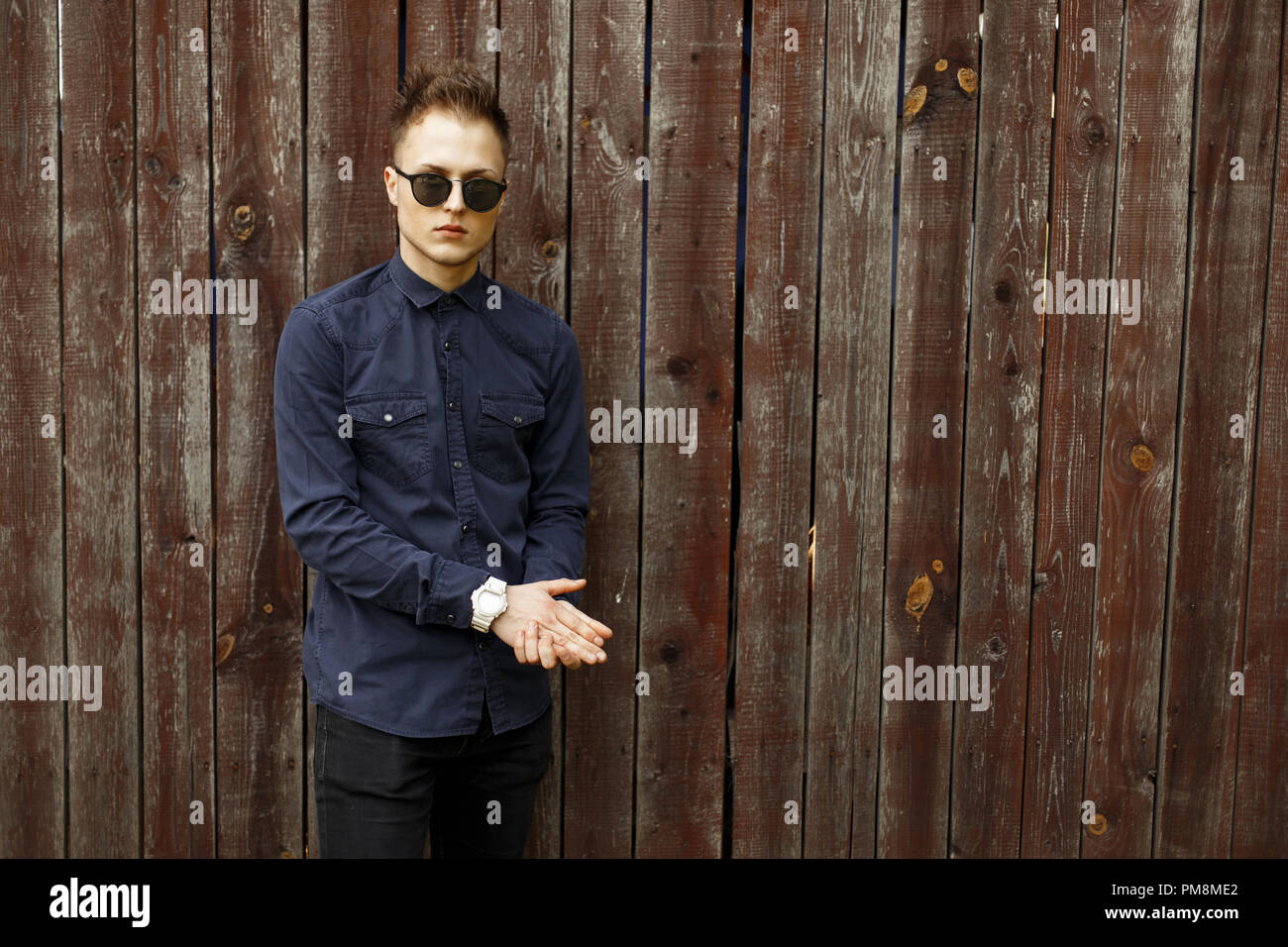 Man in fashion sunglasses with a blue shirt and black pants near a vintage wooden wall Stock Photo