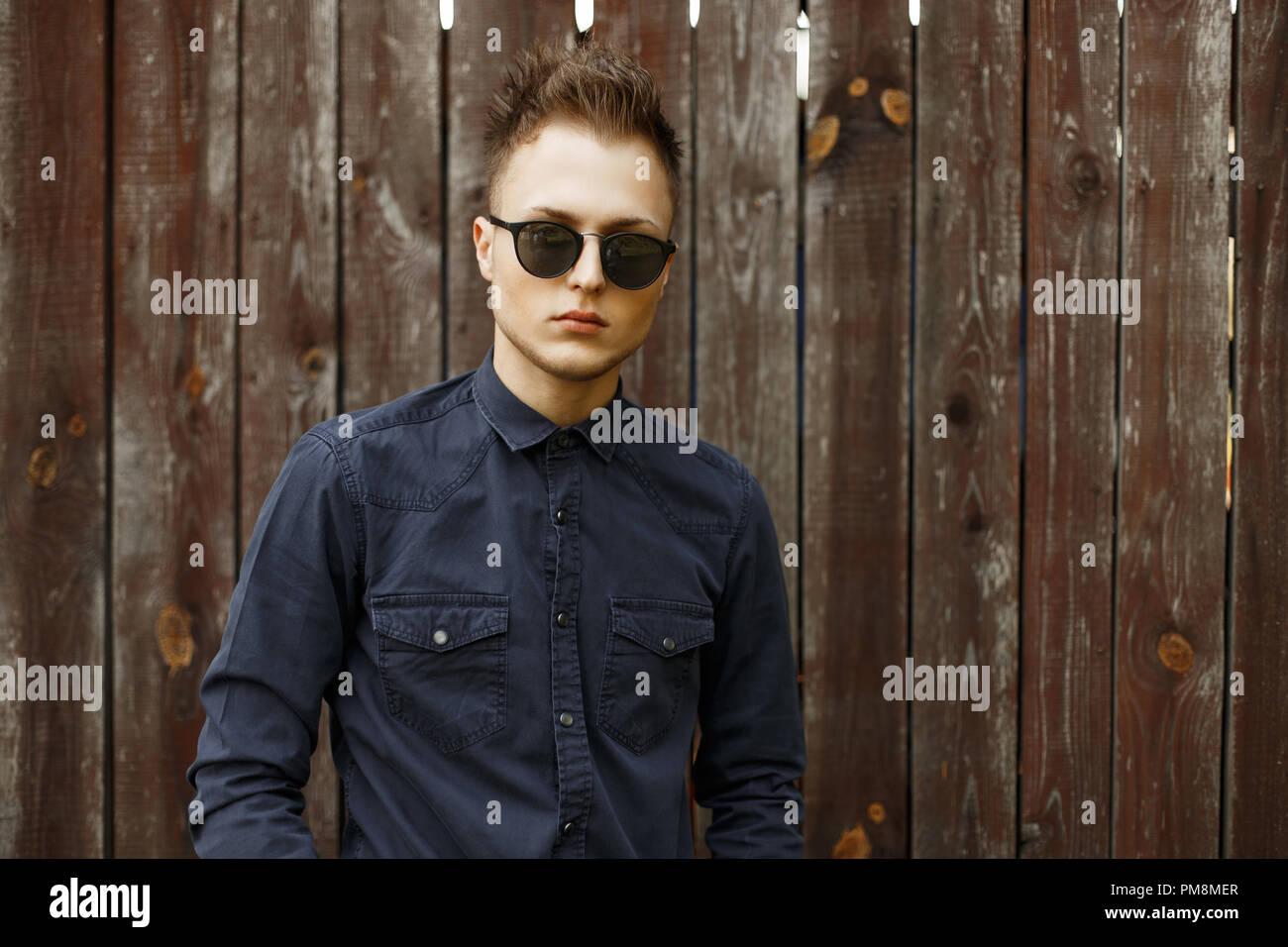 Vintage portrait of a young man in sunglasses and a blue shirt near an old wooden wall Stock Photo