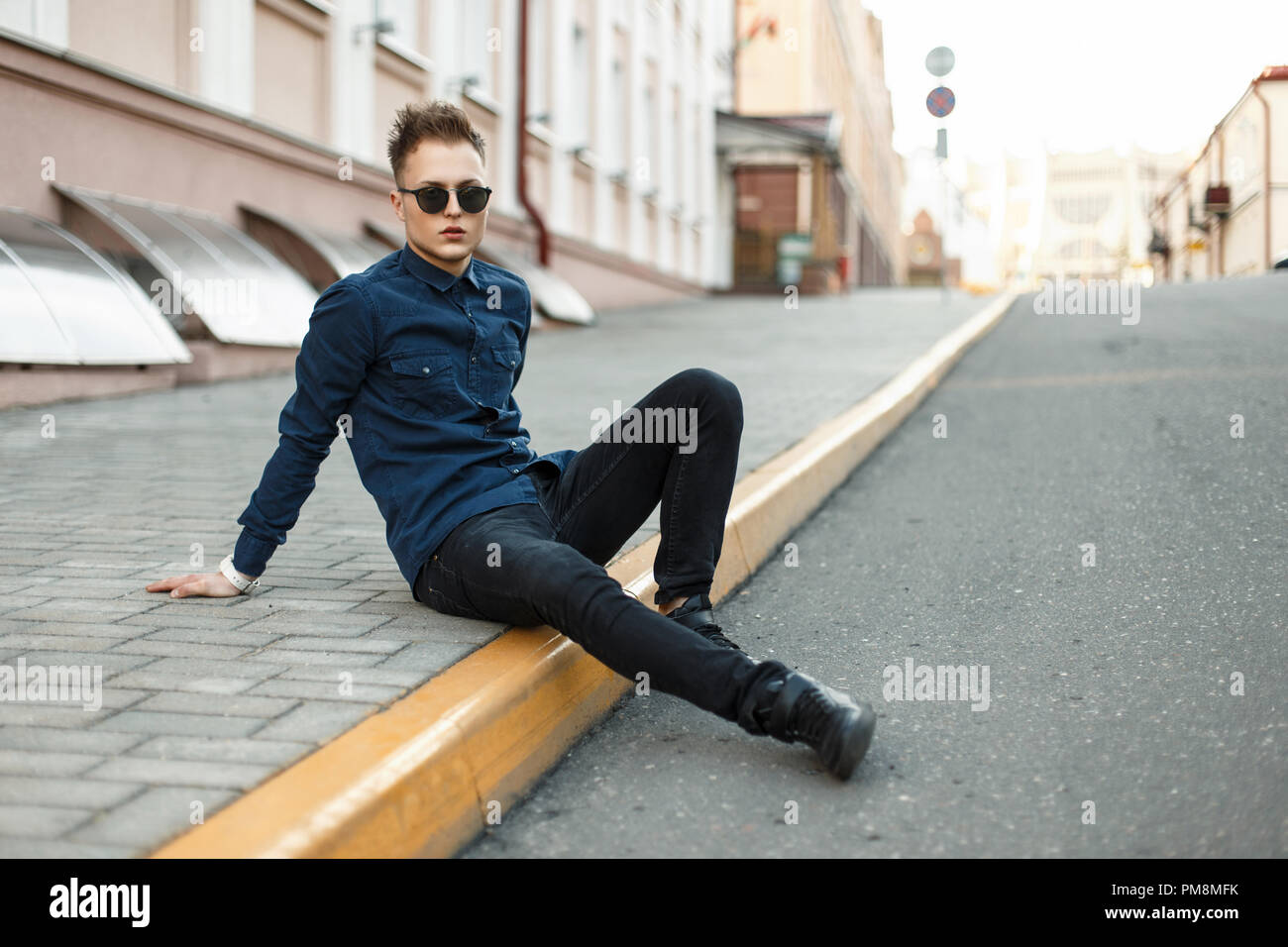 Handsome young man in a blue shirt, black jeans and sneakers sits on the sidewalk Stock Photo