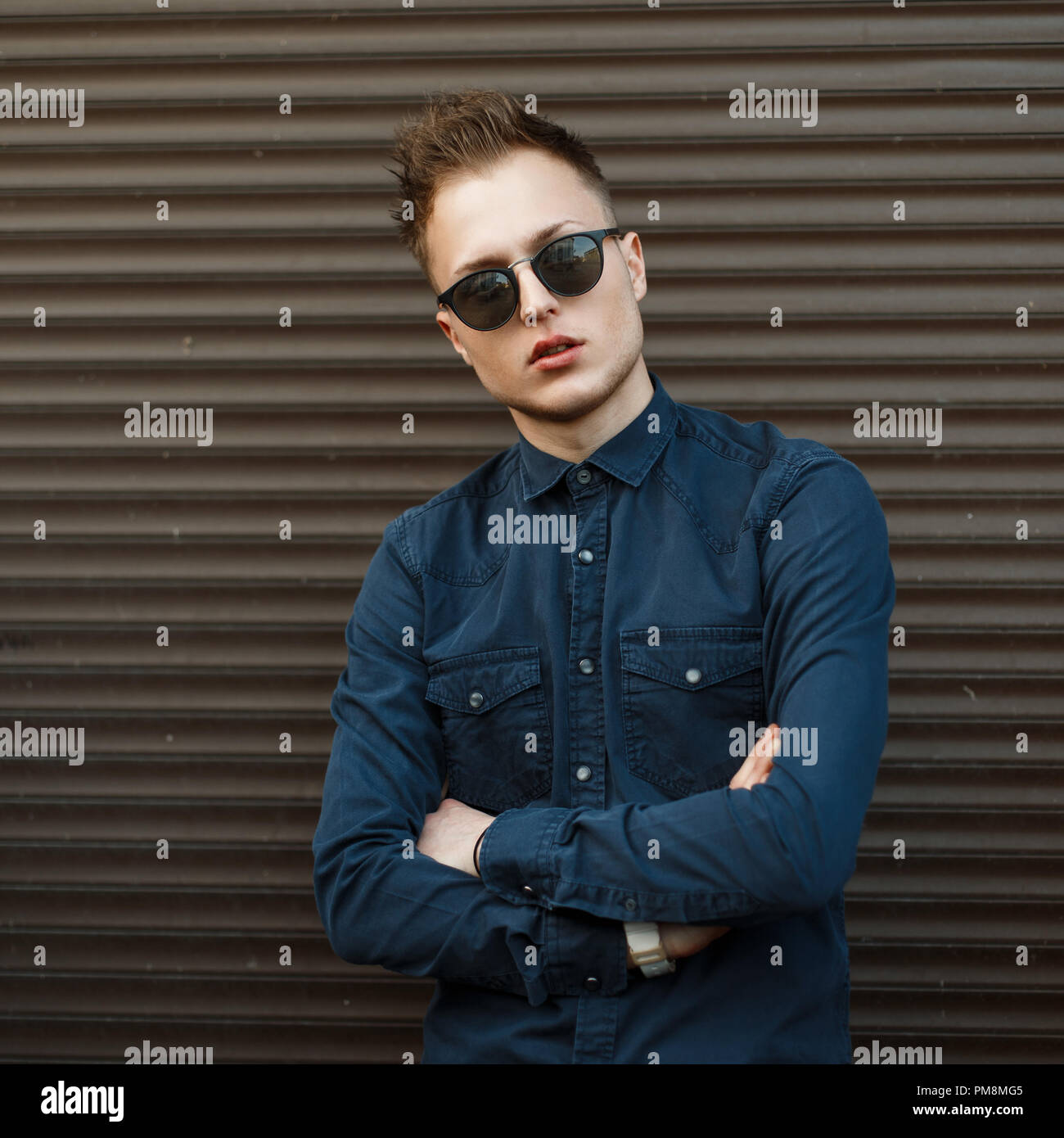 Handsome young man in a blue shirt and sunglasses near a metal wall Stock Photo