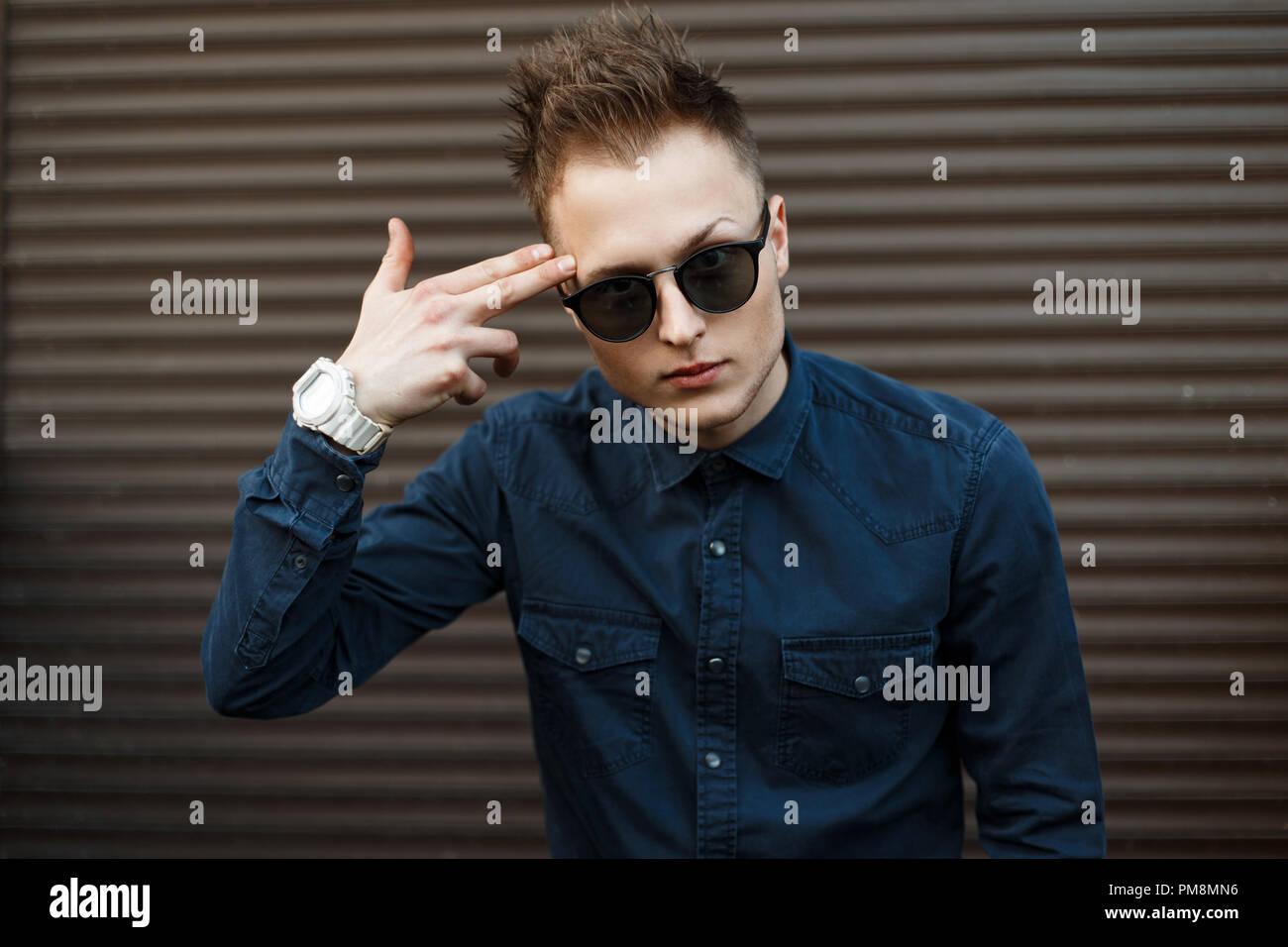 Handsome young man in sunglasses and a blue shirt shows a pistol from his fingers near the metal wall Stock Photo