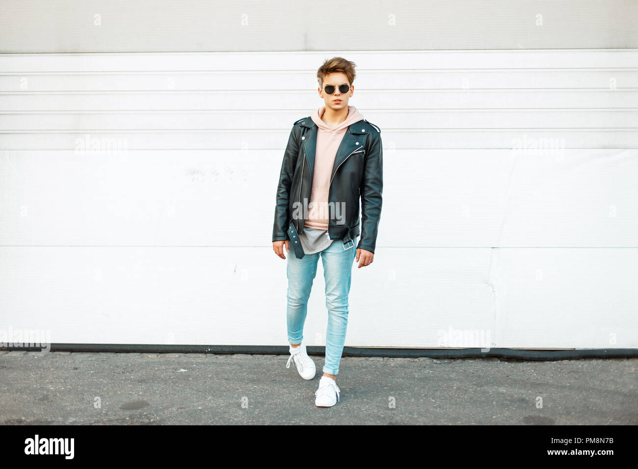 Stylish handsome young man with sunglasses in a black leather jacket, blue fashionable jeans and white trendy sneakers near a white metal wall Stock Photo