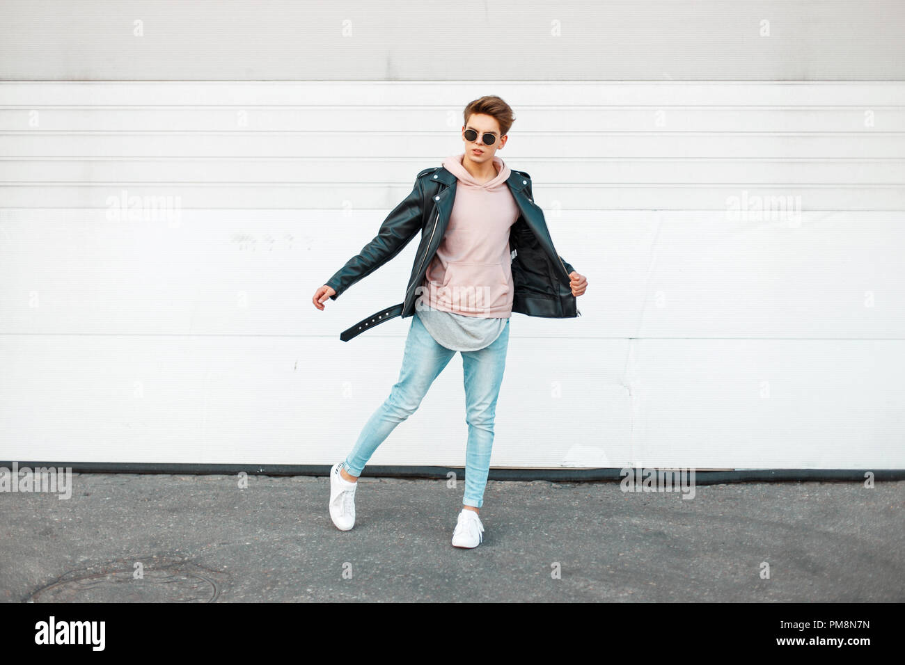 Handsome young fashionable man with sunglasses in a black leather jacket, a pink sweatshirt, blue brand jeans and white shoes in motion Stock Photo