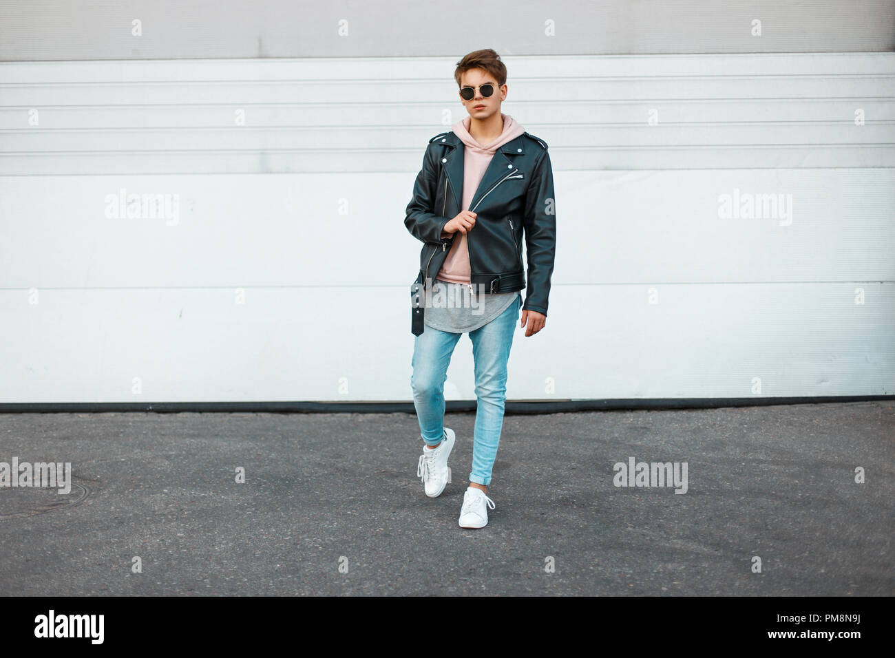 Handsome young model of a man with sunglasses in a black leather jacket, a pink sweatshirt, blue jeans and white sneakers near a metal white gate Stock Photo