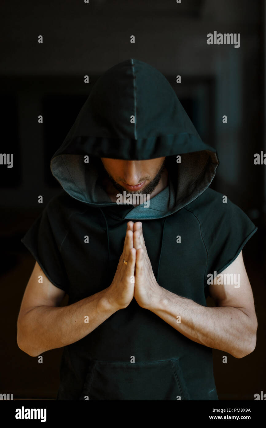 Sporty yoga man in a hood holds his hands on a dark background Stock Photo