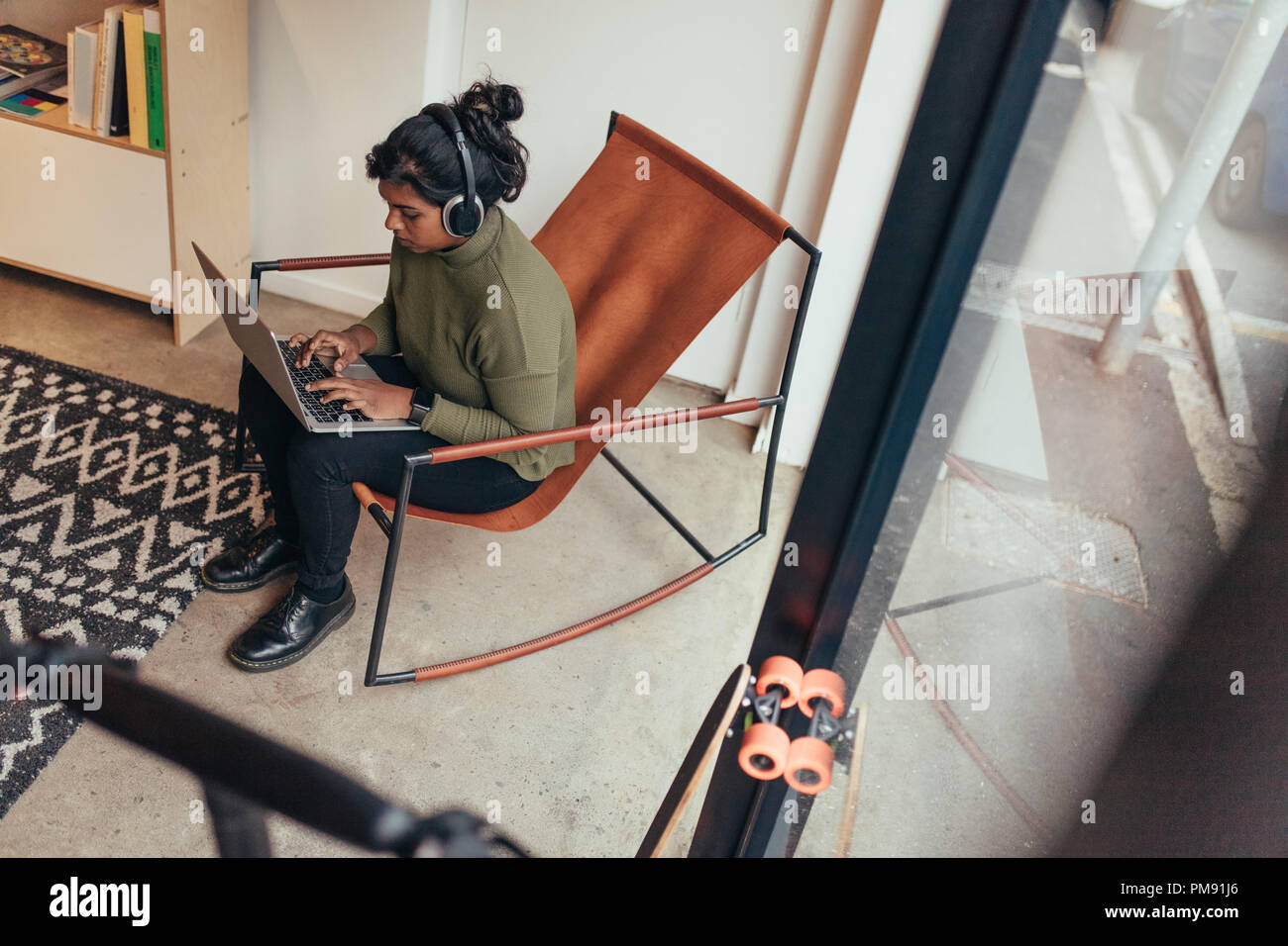 Young woman wearing headphones sitting on chair and working on laptop computer. Female computer programmer working at her startup office. Stock Photo