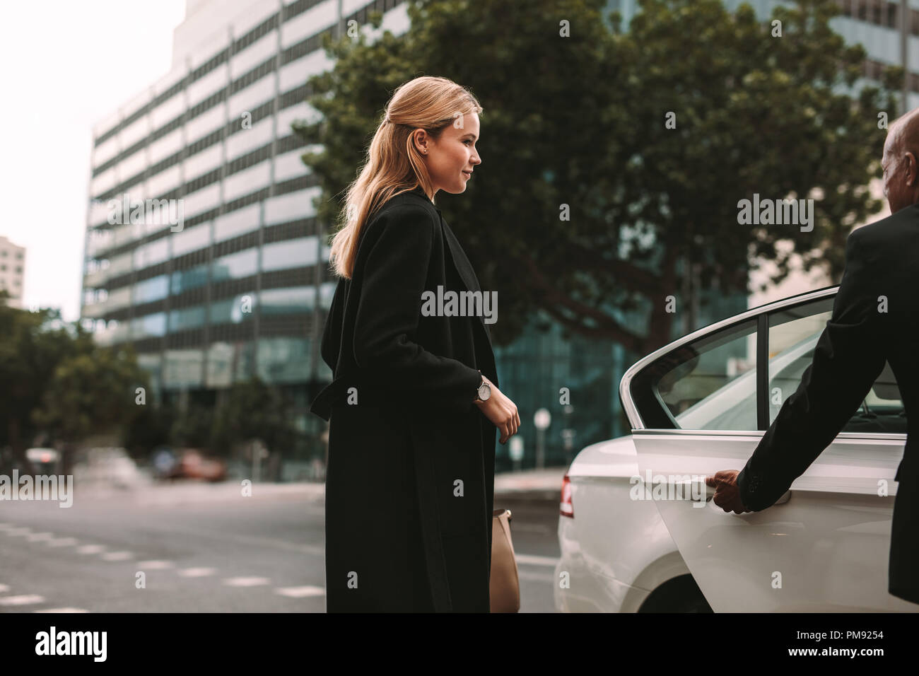 Young female commuter getting into a taxi. Businesswoman entering a taxi with driver opening door. Stock Photo