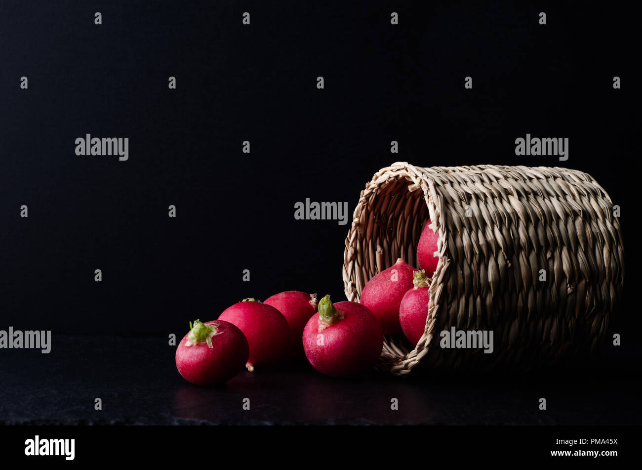 Basket filled with radishes on its side and spilling out on to slate surface with black background. Stock Photo