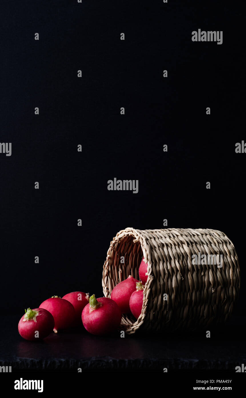 Side view of a small basket of radishes, tipped over and spilling on to slate surface with black background. Stock Photo