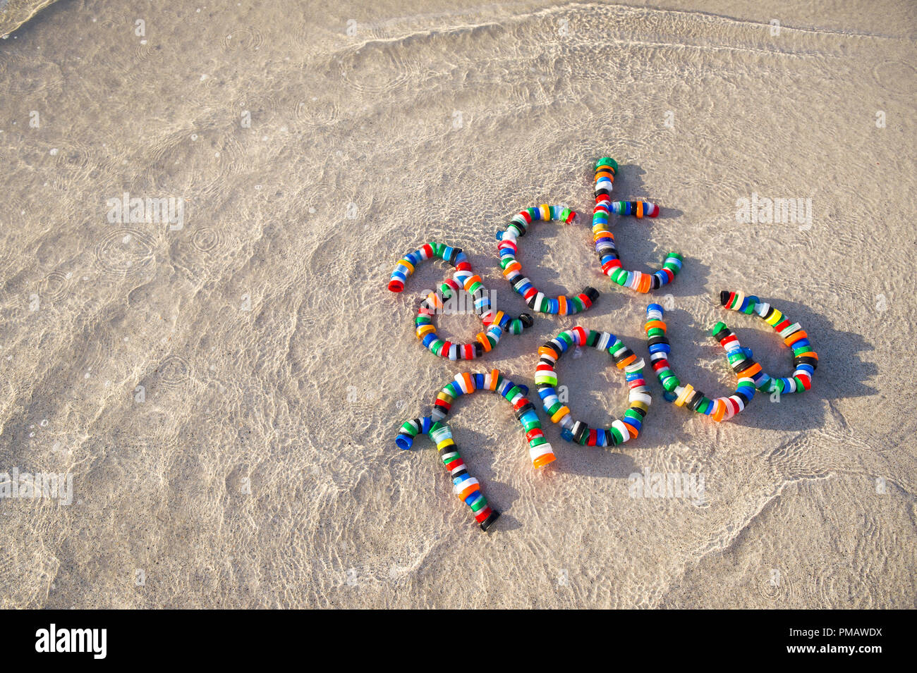 Colorful plastic bottle caps spell out 'act now' on a sandy beach as a wave laps around. A reminder for people to take action on pollution - reduce, r Stock Photo