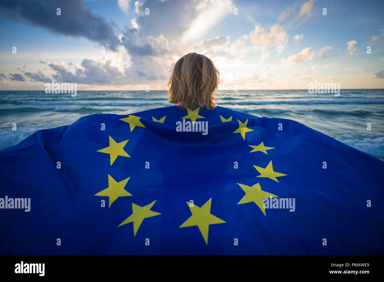 Man holding a fluttering iconic EU flag with circle of stars on beach with stormy turbulent seas in the channel at sunrise Stock Photo