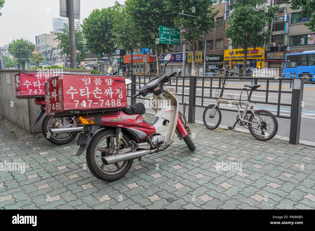 Two delivery scooters with red boxes on top, on a city street, in Seoul