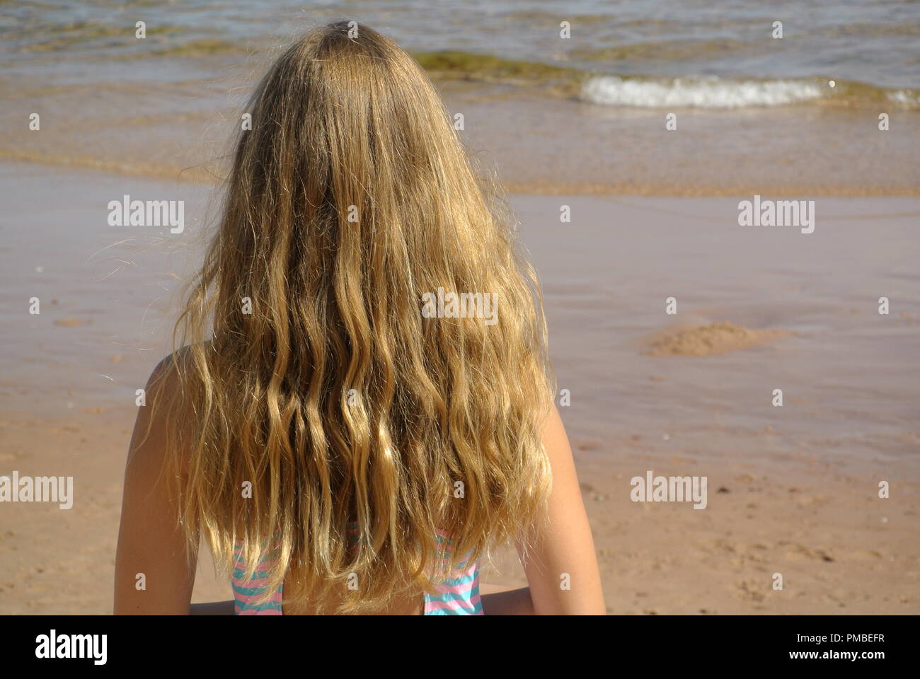 A close-up of a beautiful young girl with long blond and wavy beach hair standing on a beach and looking at the ocean on a sunny day, back view Stock Photo