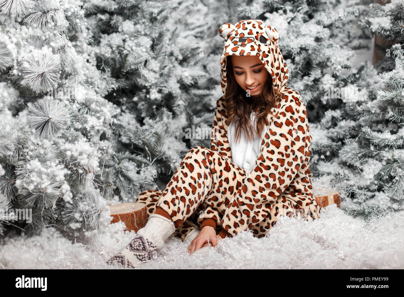 Funny young happy woman in fashionable bear wearing pajamas with hood sitting near Christmas trees with snow in studio Stock Photo