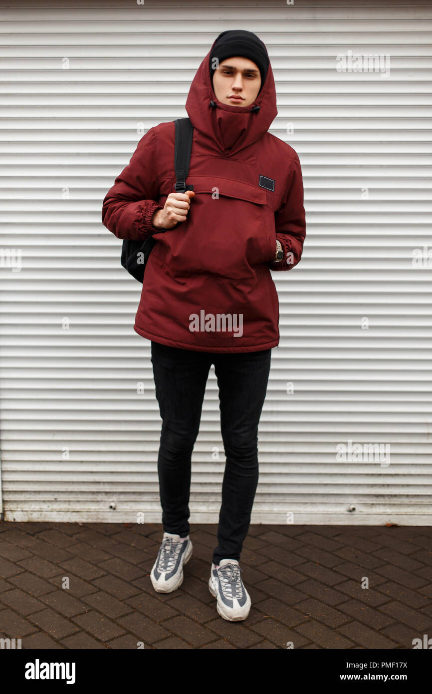 handsome young man in fashion winter sports clothes with a hat and sneakers with a backpack near white roller blinds on the street Stock Photo