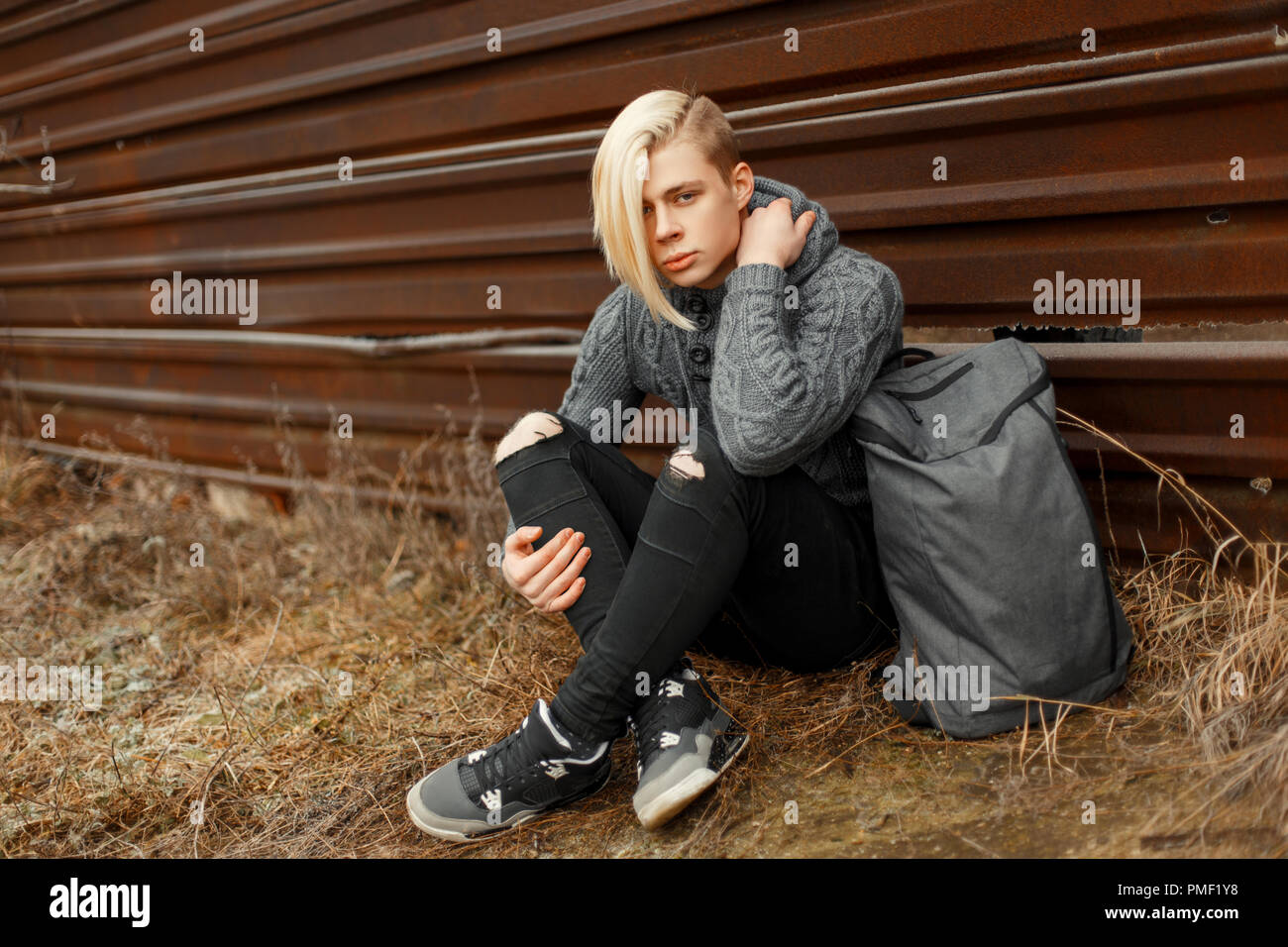 handsome young man with a haircut in a fashionable vintage knitted sweater  with black jeans and sneakers with gray backpack sits near the metal wall  Stock Photo - Alamy