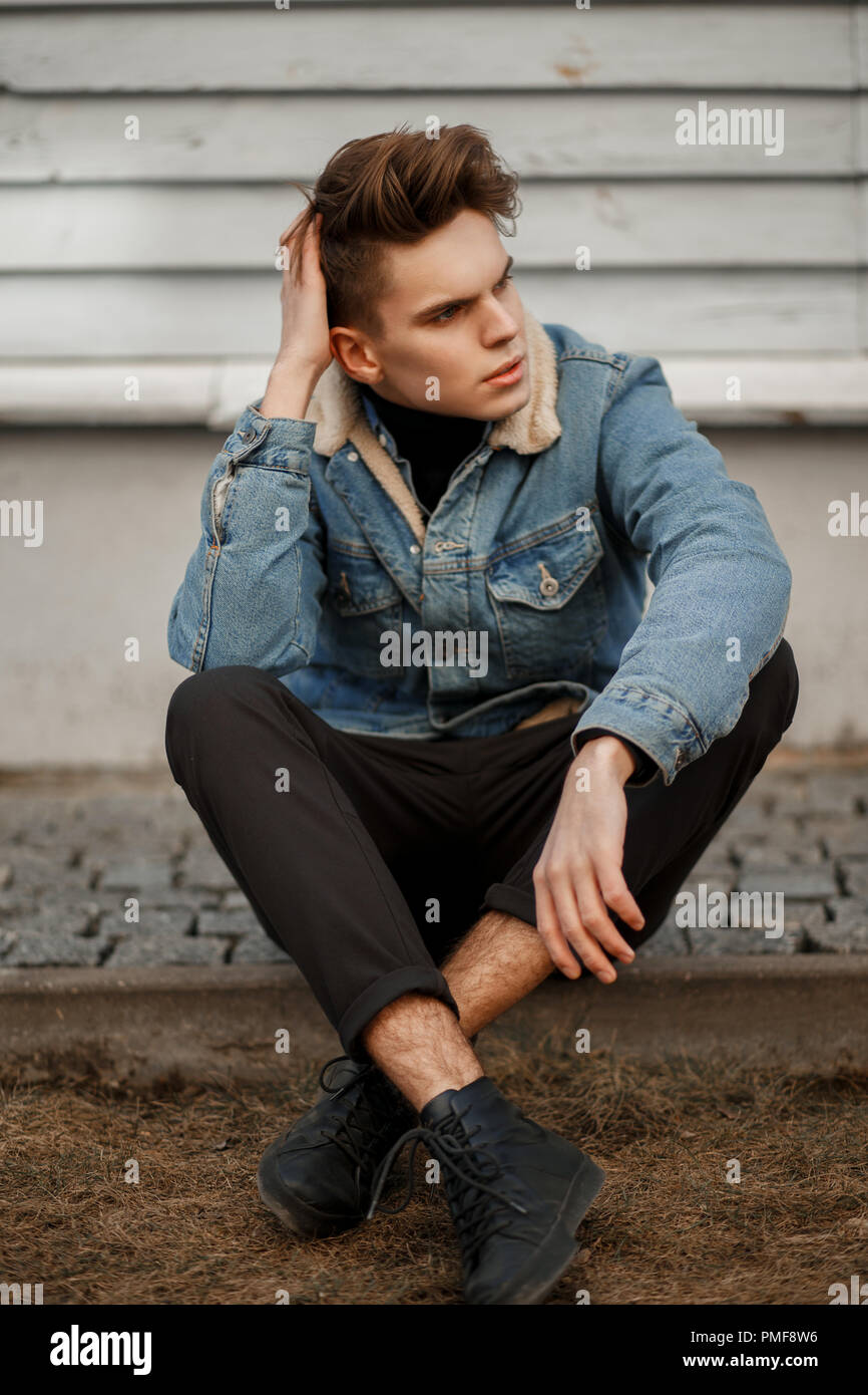 beautiful fashion model a man with a haircut in a fashionable denim jacket with black pants and shoes sitting near a wooden wall Stock Photo