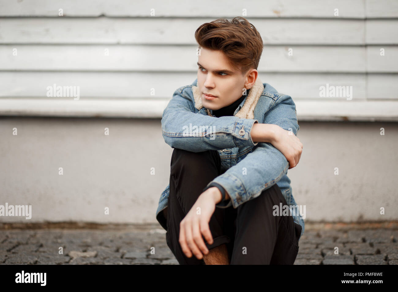 young man in fashionable denim clothes sits near a wooden wall Stock Photo
