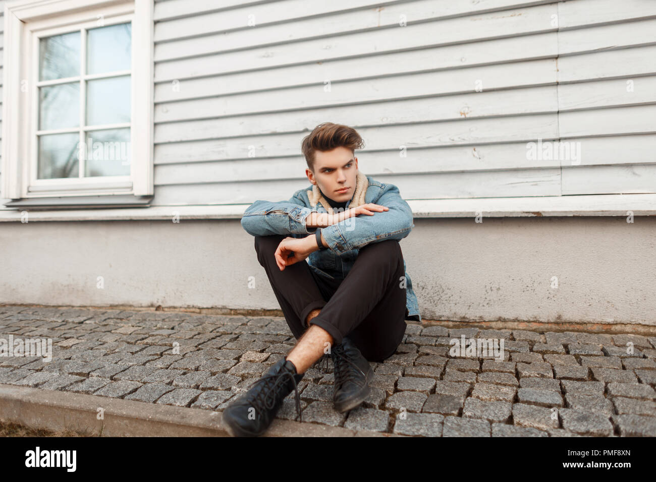 stylish fashionable young guy in a denim fashion jacket with black pants and shoes sitting near a wooden house Stock Photo