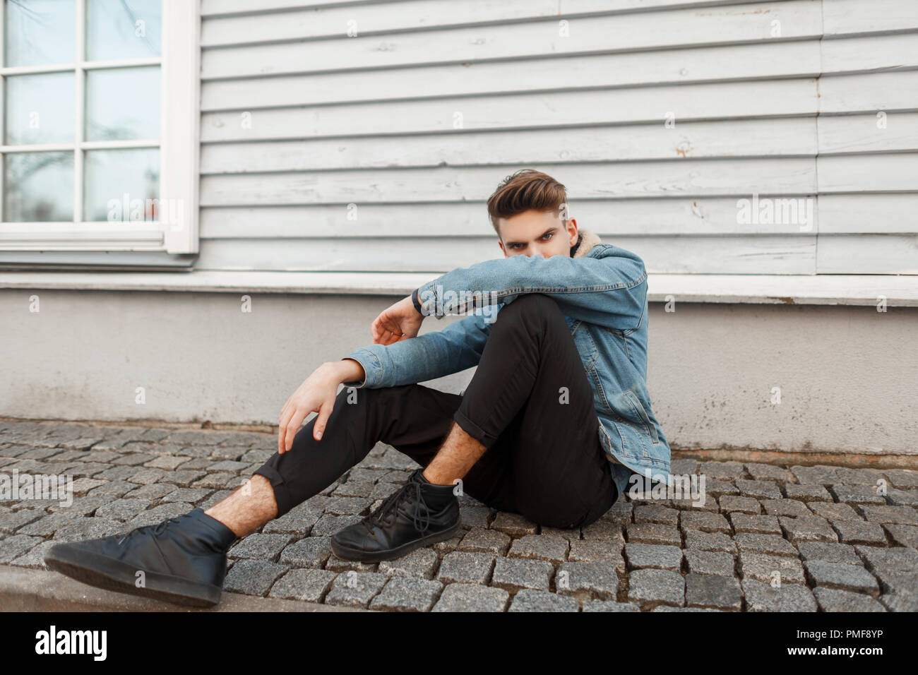 Young handsome guy model in a fashionable denim vintage jacket with black shoes sitting near a wooden house Stock Photo