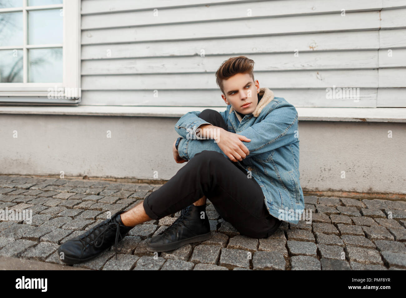 handsome stylish young man with a haircut in a fashionable vintage jeans jacket with black sneakers sits near a wooden wall Stock Photo