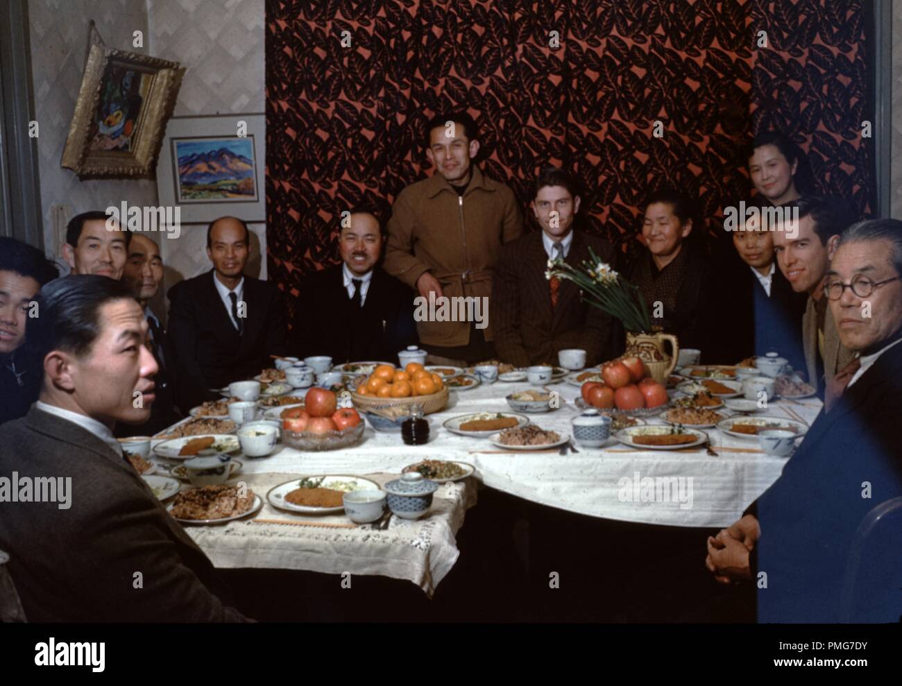 A large group of people, including Caucasian Christian missionaries and Japanese congregants, sit at a table in a small room set with a lavish meal with both Western and Japanese elements, including noodles, apples, and bowls of soy sauce, Japan, 1955. () Stock Photo