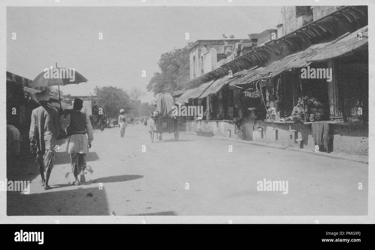 Black and white photograph on cardstock, of a dusty road, with awning-covered, terraced buildings at either side suggesting a market street, with two dhoti-clad men (one holding an umbrella over the other's head) walking in the left foreground, a cart at mid-ground, and a turban-clad man standing in the middle of the road in the background, likely collected as a tourist souvenir during a trip to South Asia (likely India), 1910. () Stock Photo