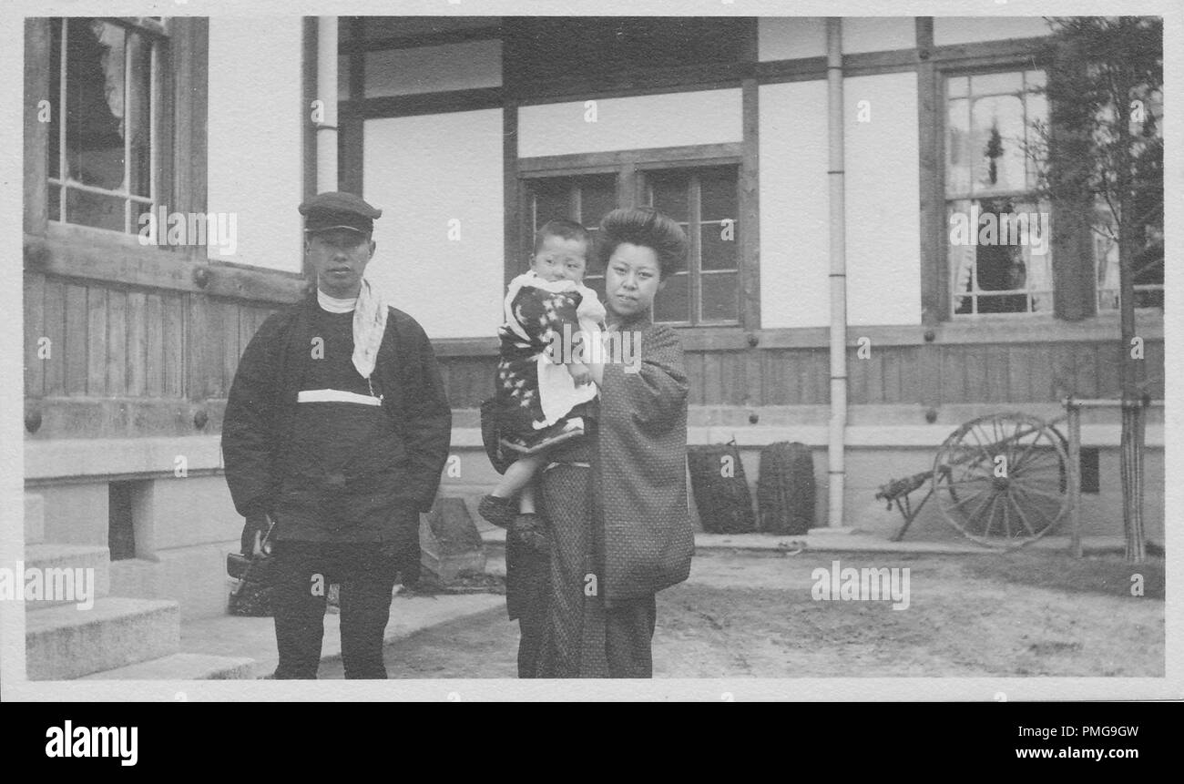 Black and white photograph on cardstock, of a Japanese man and woman (the latter holding a small child) facing the camera, with serious expressions on their faces, each wearing circa 1900s, Meiji-era clothing, the man in a woolen cap, jacket, and trousers ensemble, the woman and child wearing yukatas or kimonos, with a large, wooden and stone structure visible in the background, likely collected as a tourist souvenir during a trip to Japan, 1910. () Stock Photo