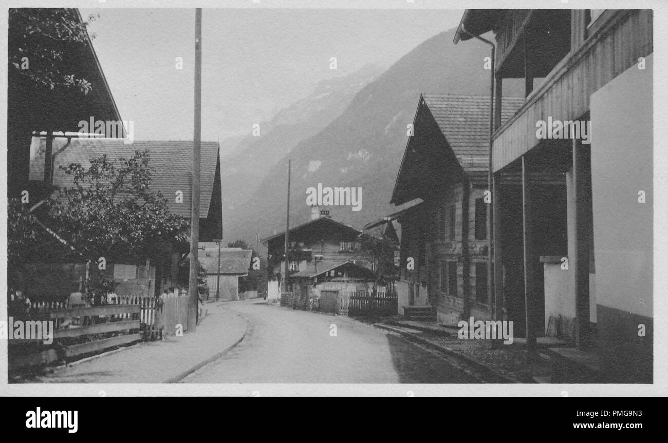 Black and white photograph on cardstock, with an image of a rural, European street, with multi-story, wooden chalets, and with mountains, possibly the Alps, looming in the background, likely collected as a tourist souvenir during a trip to Northern Europe (likely a region of the Upper Rhine, near the borders of Germany, Switzerland, and France), 1910. () Stock Photo