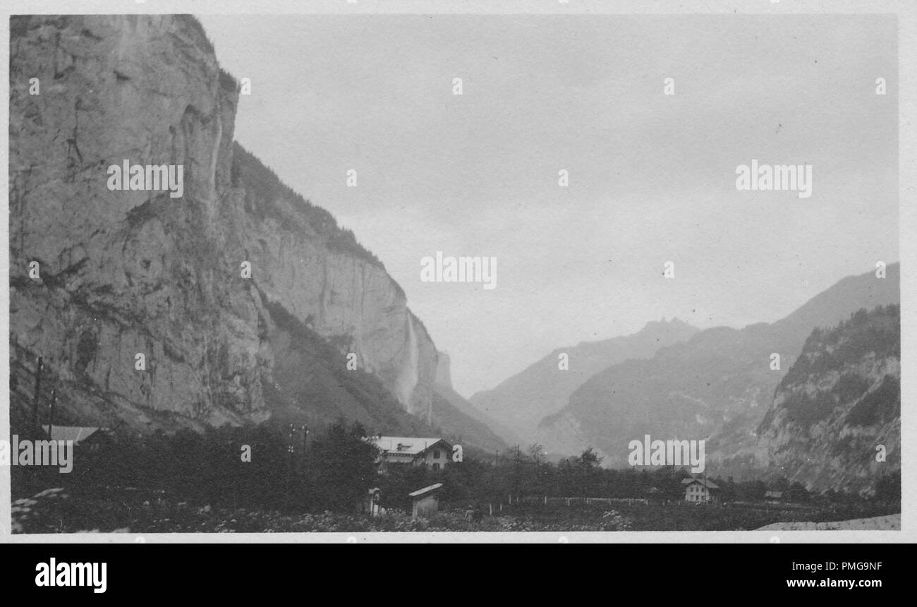 Black and white photograph on cardstock, with an image of a several multi-story, wooden chalets, nestled amidst trees in a rural setting, with the stark cliff faces of large, rocky mountains (possibly the Alps) looming in the background, likely collected as a tourist souvenir during a trip to Northern Europe (likely a region of the Upper Rhine, near the borders of Germany, Switzerland, and France), 1910. () Stock Photo