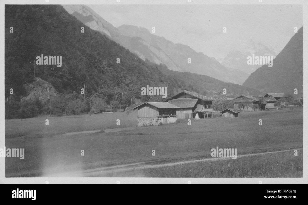 Black and white photograph on cardstock, with an image of a several multi-story, wooden chalets, in a grassy, rural setting, with tree-covered and snow-capped mountains (possibly the Alps) looming in the background, likely collected as a tourist souvenir during a trip to Northern Europe (likely a region of the Upper Rhine, near the borders of Germany, Switzerland, and France), 1910. () Stock Photo