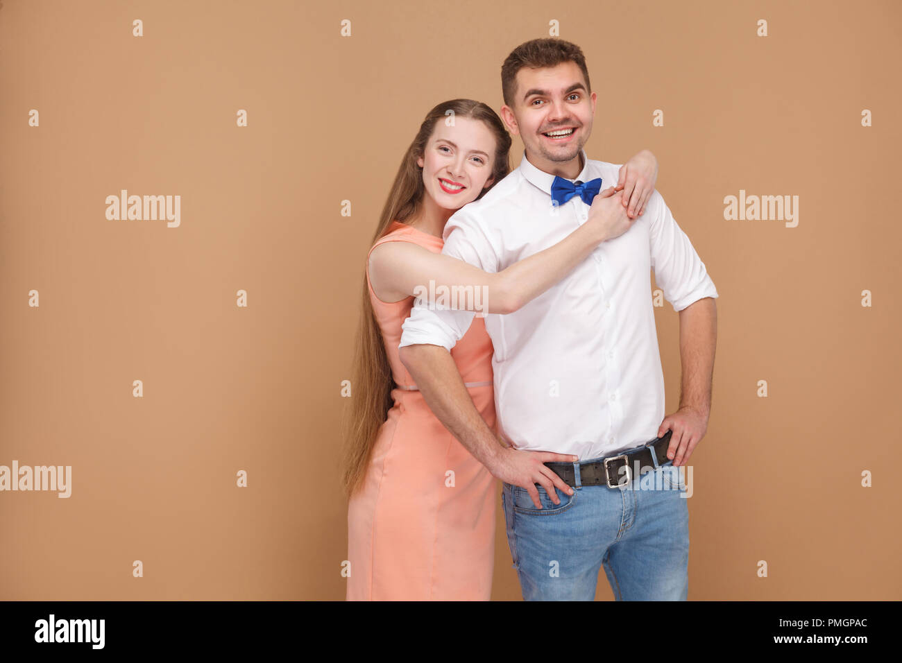 Portrait of handsome man in casual style and beautiful woman in pink dress or happy couple, hugging and looking at camera with toothy smile. indoor st Stock Photo