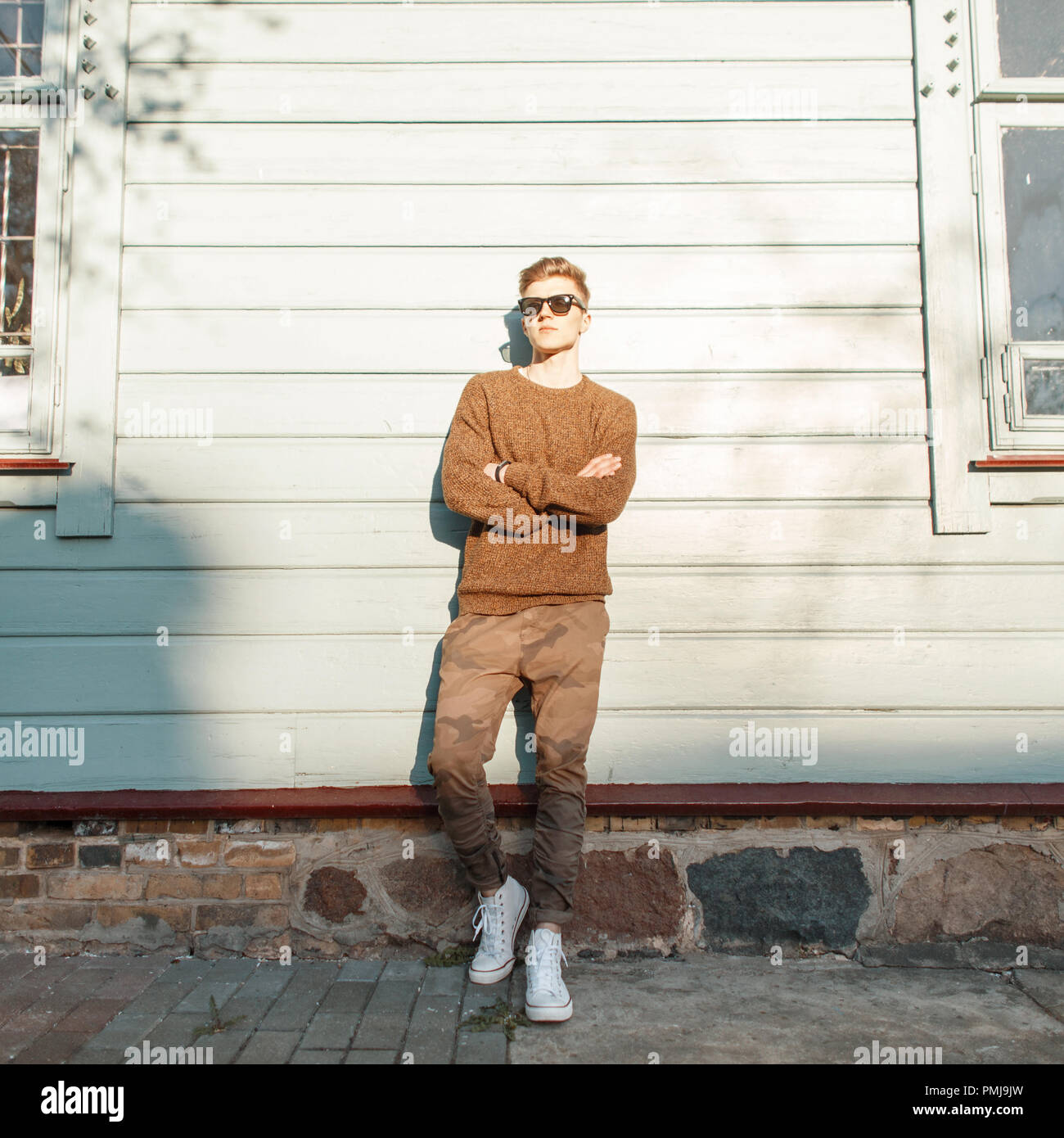 Fashionable young hipster man model with sunglasses in fashionable military pants with white shoes posing near a wooden vintage wall at sunset Stock Photo