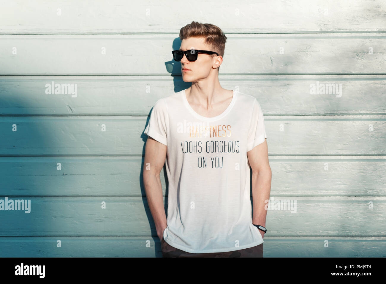Handsome fashion man hipster in stylish sunglasses in a white T-shirt posing near a blue wooden wall on the beach on a sunny day. Happiness looks gorg Stock Photo