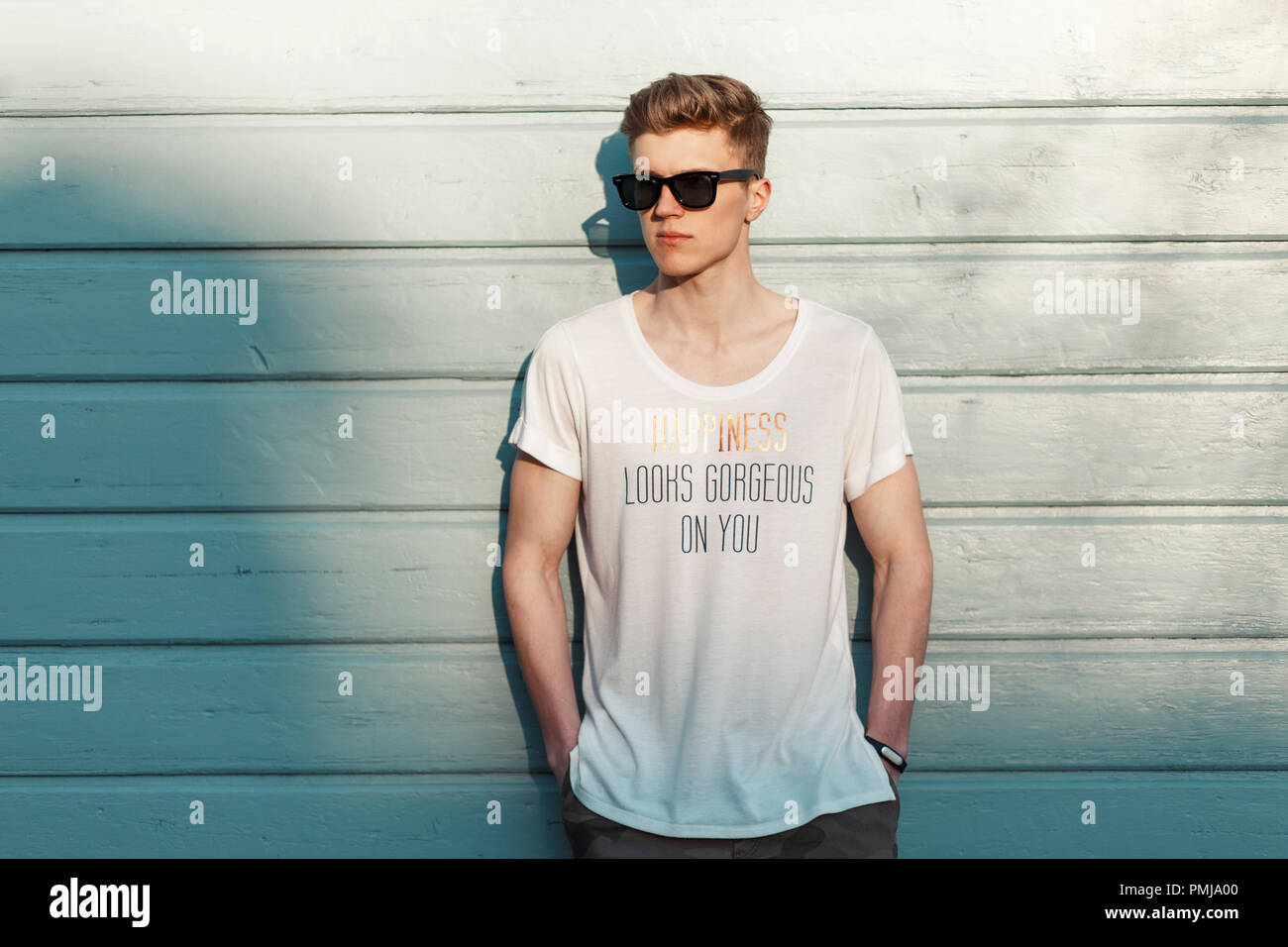 Stylish young man with sunglasses in a fashion white T-shirt posing near a blue wooden wall on the beach Stock Photo