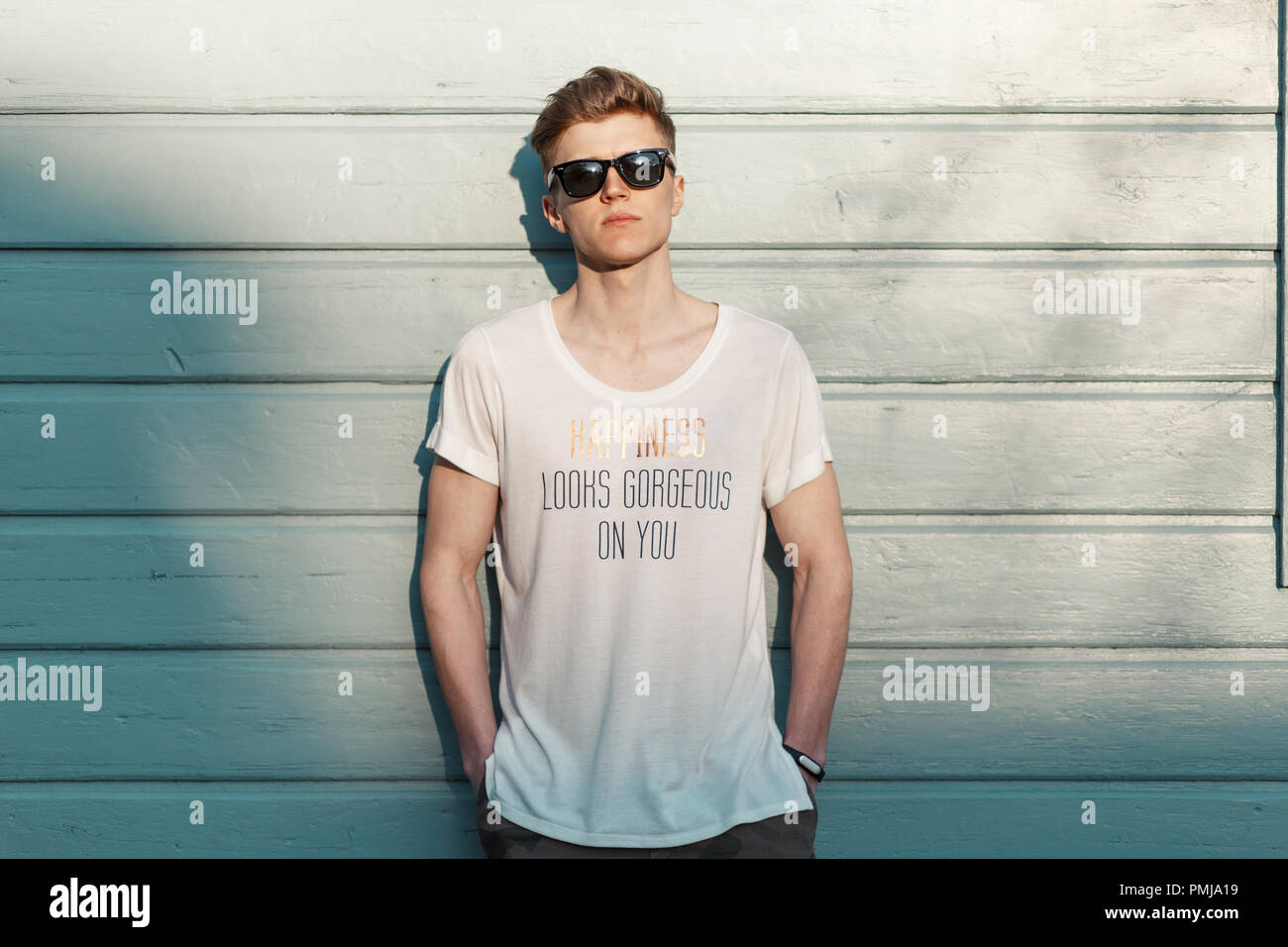 Handsome young stylish model man in sunglasses with a stylish white t-shirt stands near a wooden blue wall Stock Photo