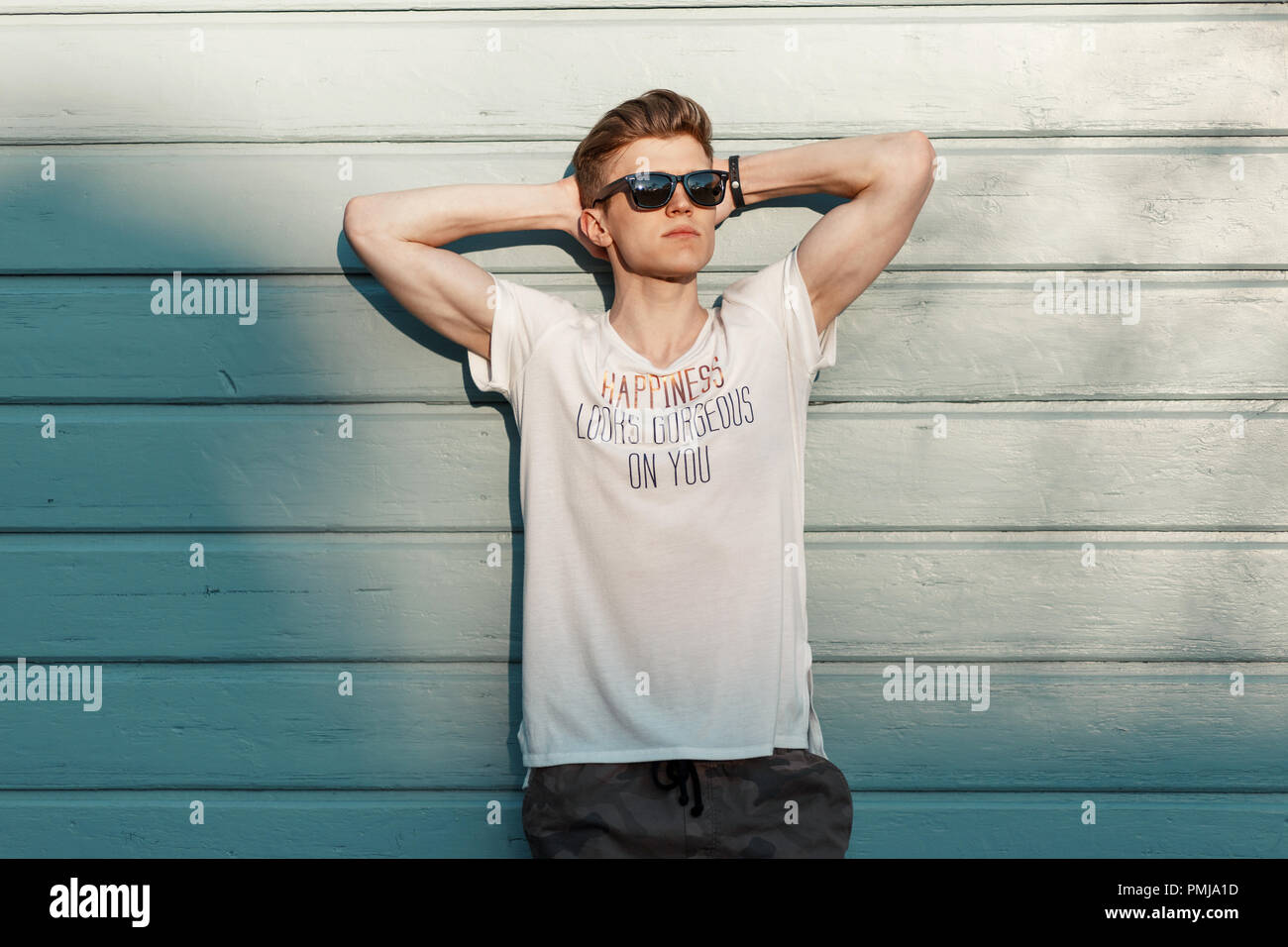 Handsome young hipster man with sunglasses in fashion clothes resting near a blue wooden beach wall on a sunny day Stock Photo