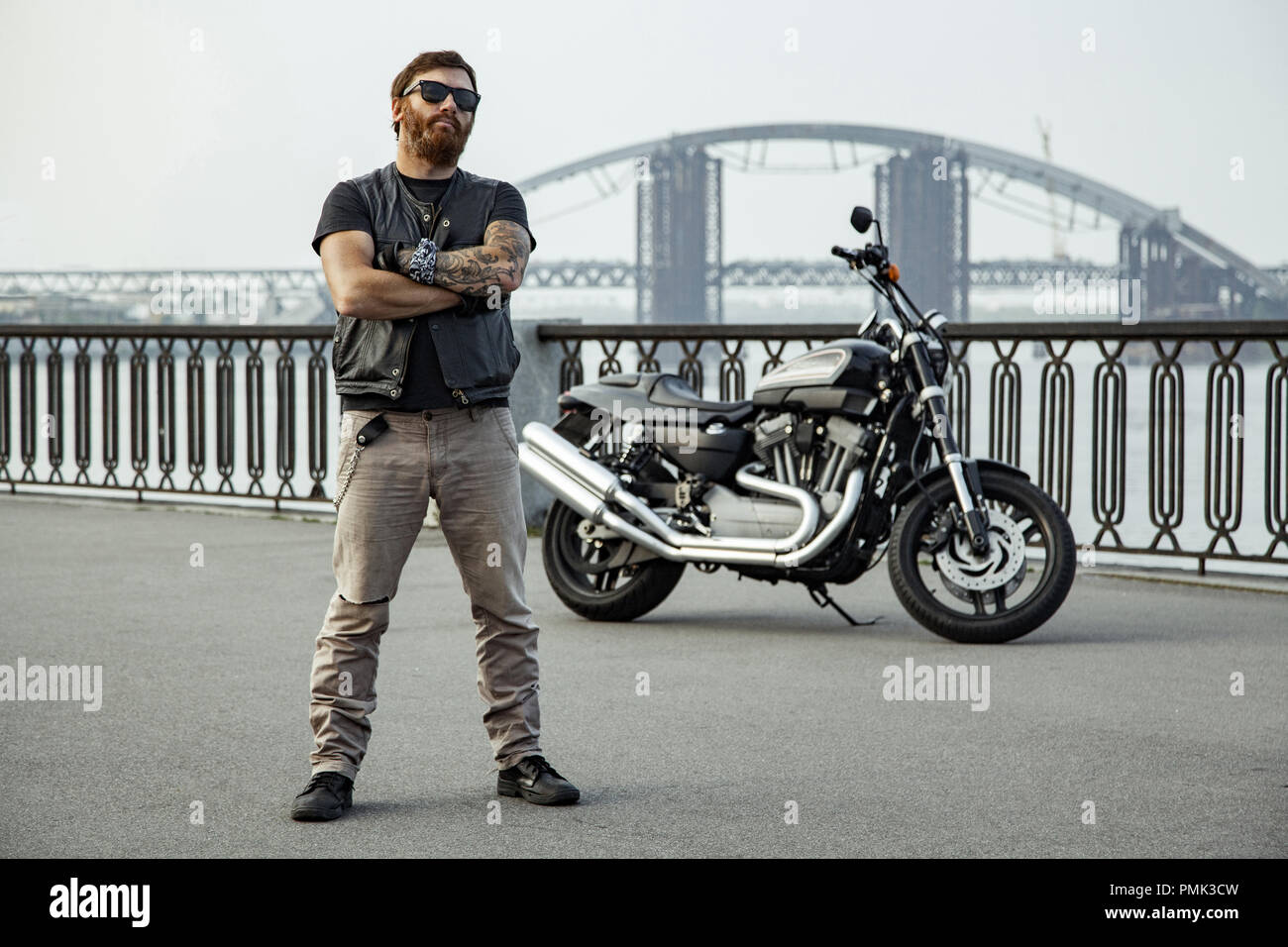 Redhead biker with beard in leather jacket standing with crossed arms near motorbike on bridge Stock Photo