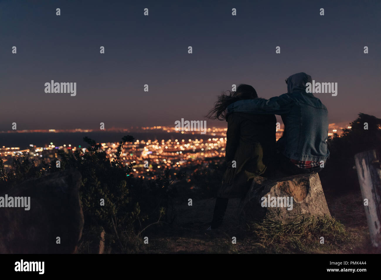 Couple enjoying the view of a brightly lit city sitting on top of a hill at night. Man sitting with a woman on a hill with his arm around her shoulder Stock Photo