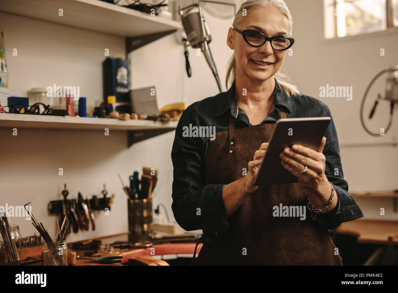 Portrait of happy mature woman jewelry maker using digital tablet in her workshop. Smiling senior woman in goldsmith workshop with tablet pc. Stock Photo