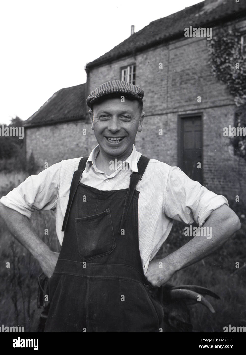 1950s, historical, portrait of a male estate maintenance man or handyman standing outside a farm property, wearing his work clothes, collared shirt, dunagarees and flat cap, England, UK. Stock Photo