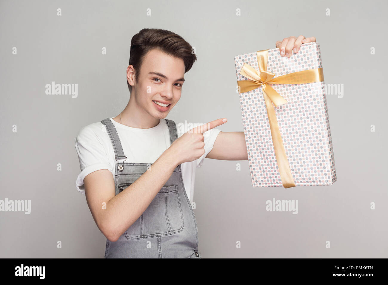 Cheerful modern teenager in demin overalls and white t-shirt standing and pointing finger to present with yellow bow and toothy smile, looking at came Stock Photo