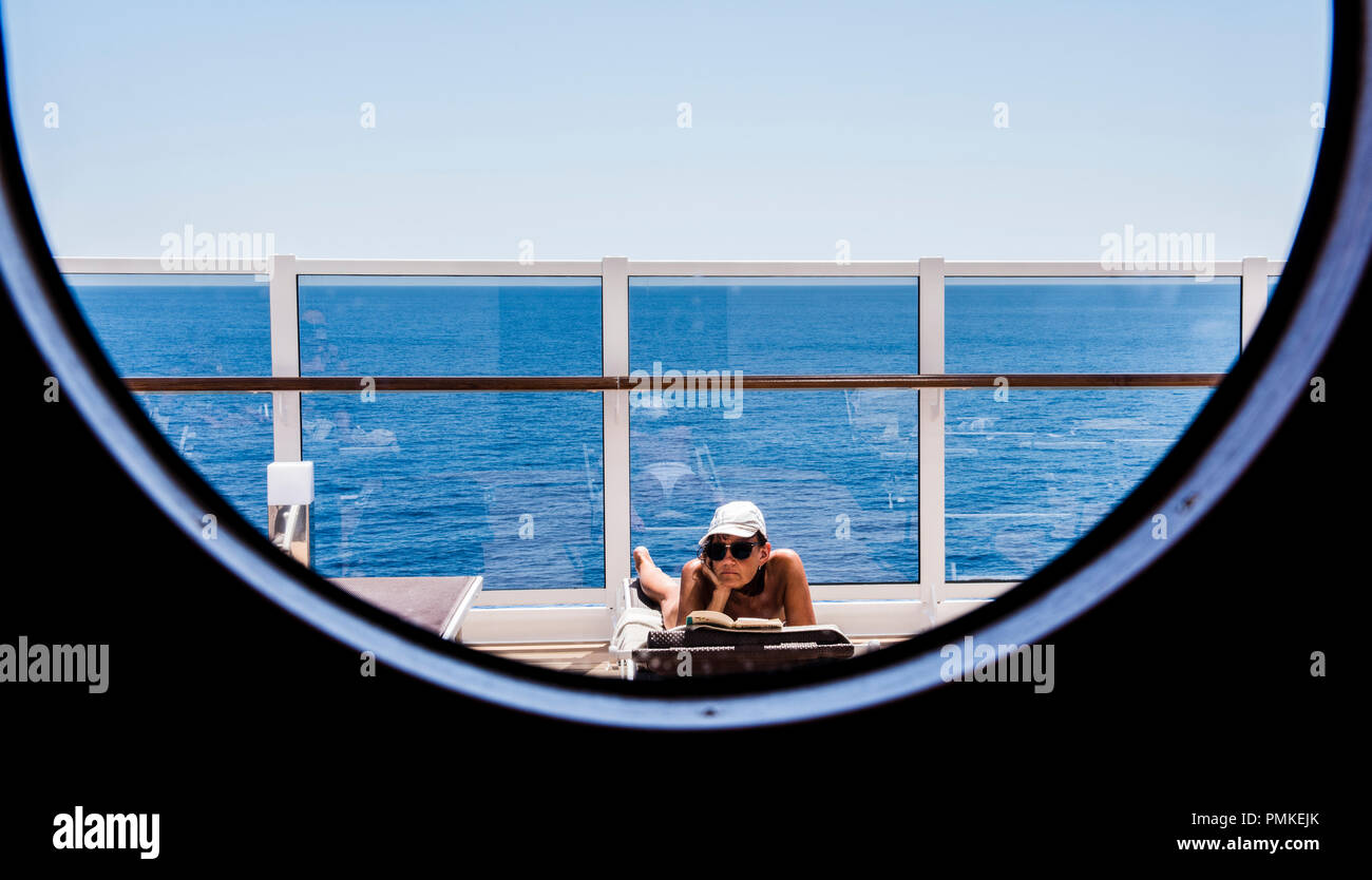 woman sunbathing and reading on deck, seen through a round ship window