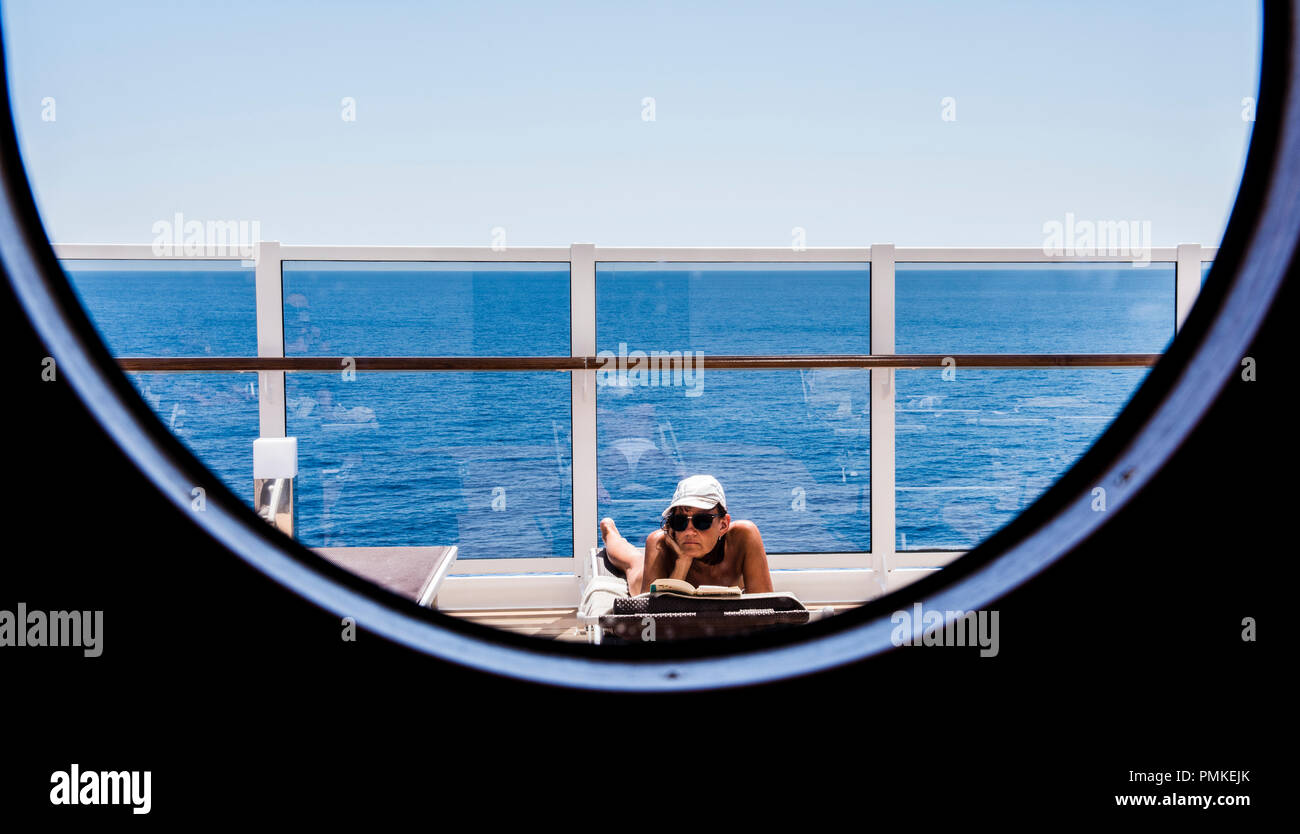 woman sunbathing and reading on deck, seen through a round ship window on cruise ship MSC Seaview Stock Photo