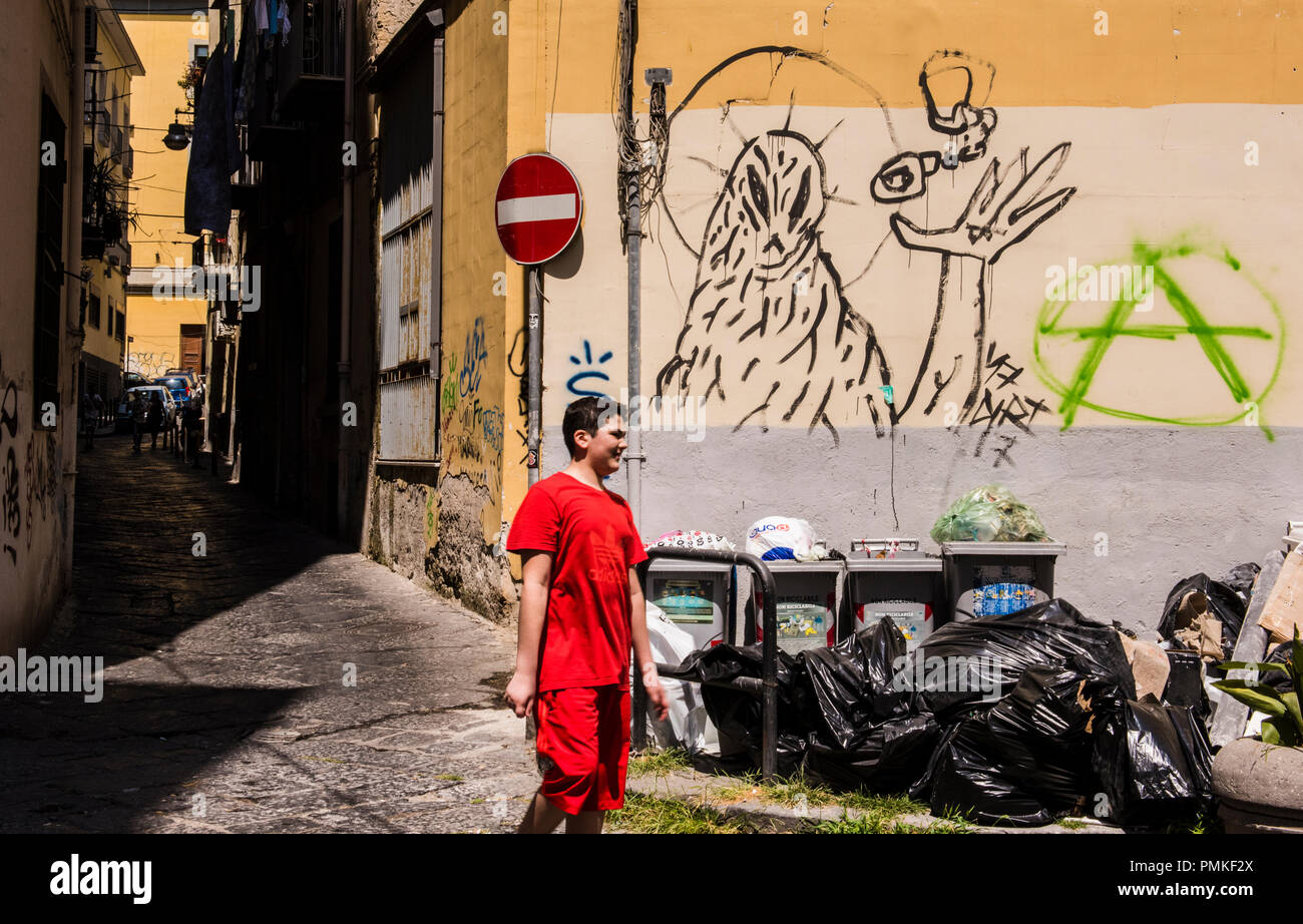 Boy dressed in red, walking past pile of rubbish, graffiti on wall in background, Naples, Italy, Europe Stock Photo