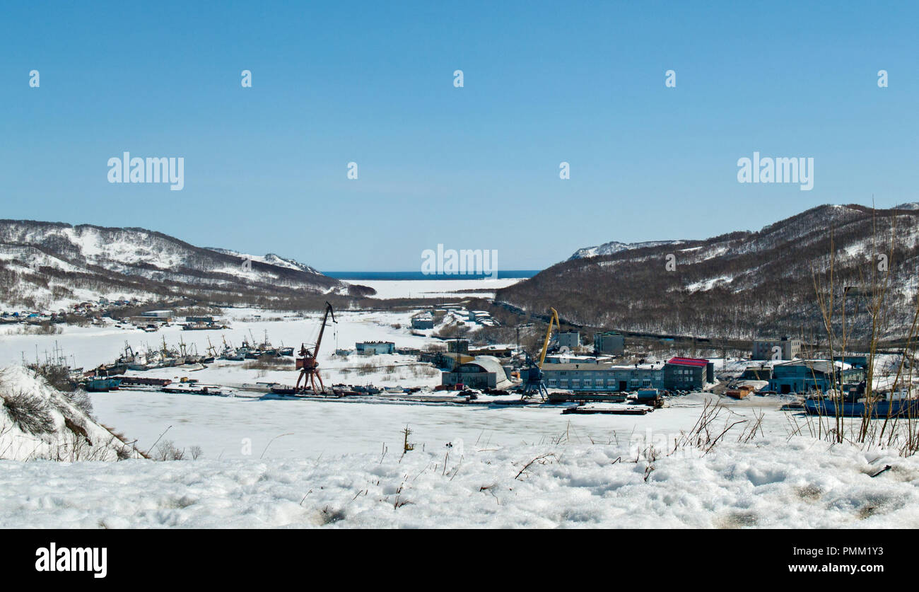 A view of a frozen ice and snow-covered bay with ships and production buildings against the background of winter hills in Petropavlovsk-Kamchatsky Stock Photo