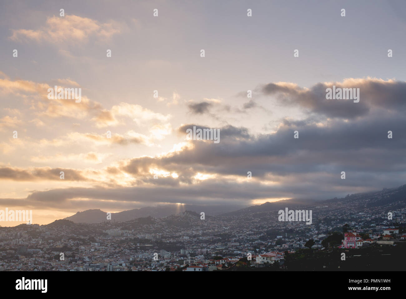 Looking down over the sprawling hillside city of Funchal  with mountains in the distance. Sunset with the sun's last rays breaking through the clouds Stock Photo