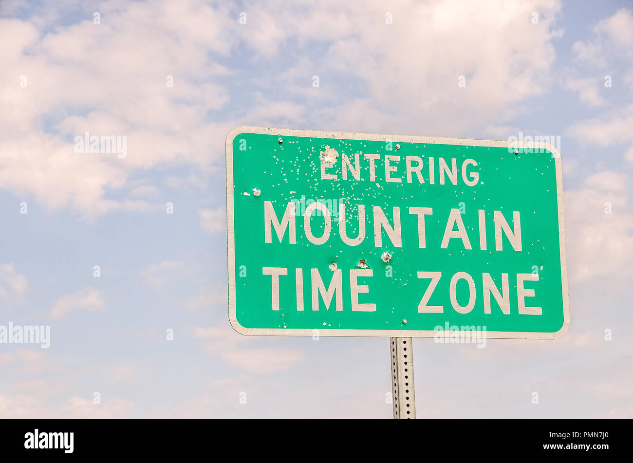 Green and white sign with bullet holes letting travelers know they are entering the mountain time zone Stock Photo